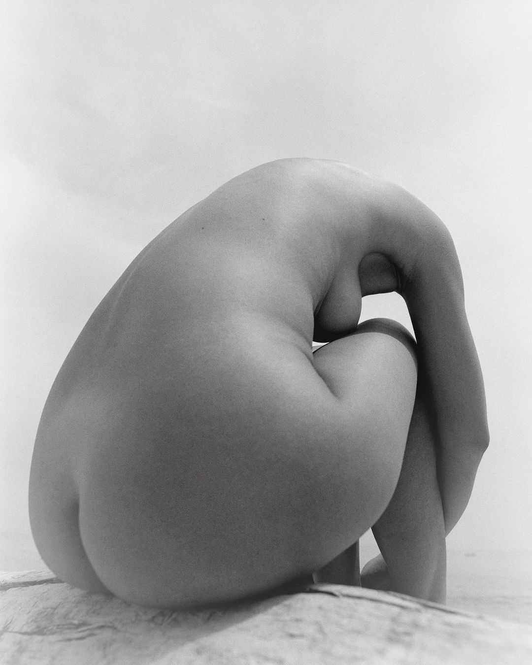Monochromatic nude art photo of a woman sitting with her head down and her arms wrapped around her legs. The figure rests on stone.