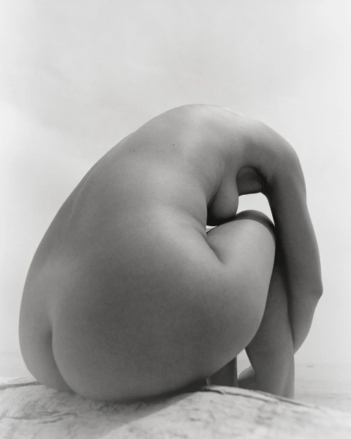 Monochromatic nude art photo of a woman sitting with her head down and her arms wrapped around her legs. The figure rests on stone.