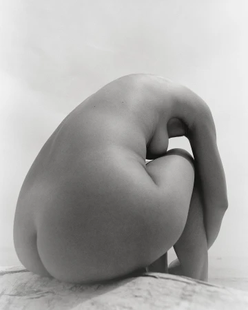 Monochromatic nude art photo of a woman sitting with her head down and her arms wrapped around her legs. The figure rests on stone.