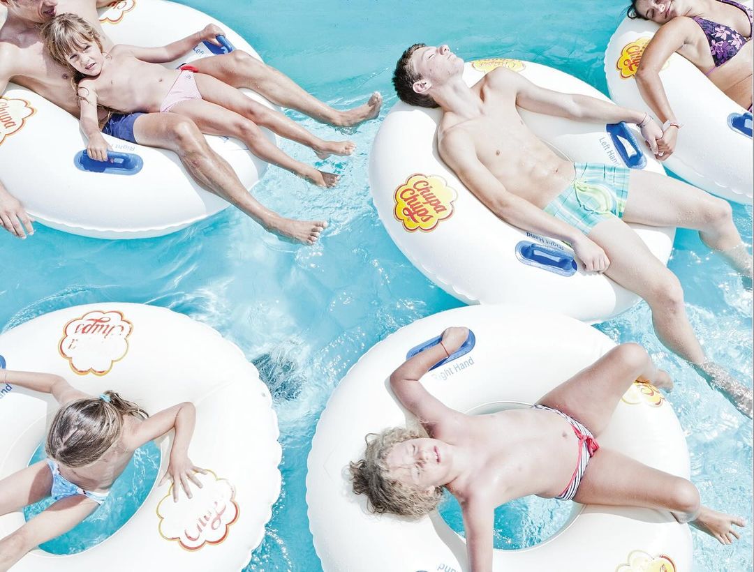 Group of adults and children relax on branded inner tubes in a sunny outdoor pool, top-down view. Light blue water reflects daylight.