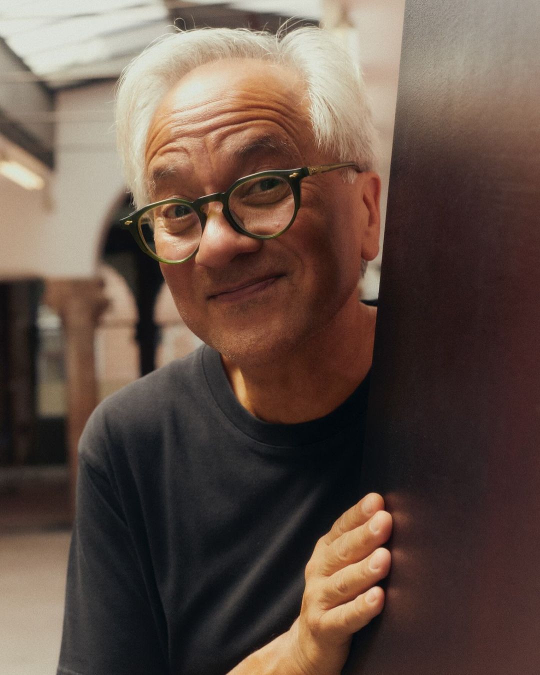 Headshot of an elderly Asian man with gray hair, wearing glasses, smiling, and leaning against a dark panel in an indoor setting.
