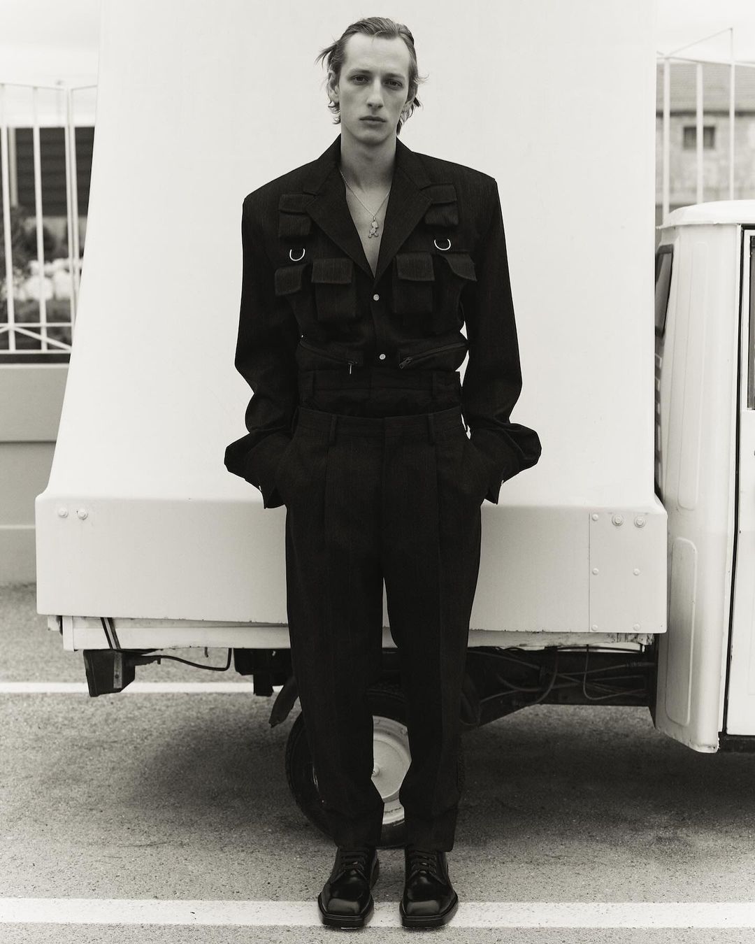 Blond male model in black jacket and trousers poses in front of white truck.  Black-and-white, fashion, outdoor, full shot.