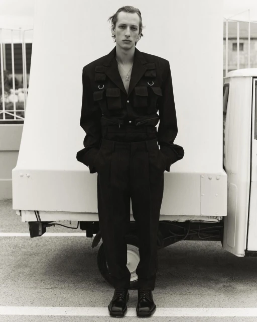 Blond male model in black jacket and trousers poses in front of white truck.  Black-and-white, fashion, outdoor, full shot.