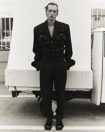 Blond male model in black jacket and trousers poses in front of white truck.  Black-and-white, fashion, outdoor, full shot.