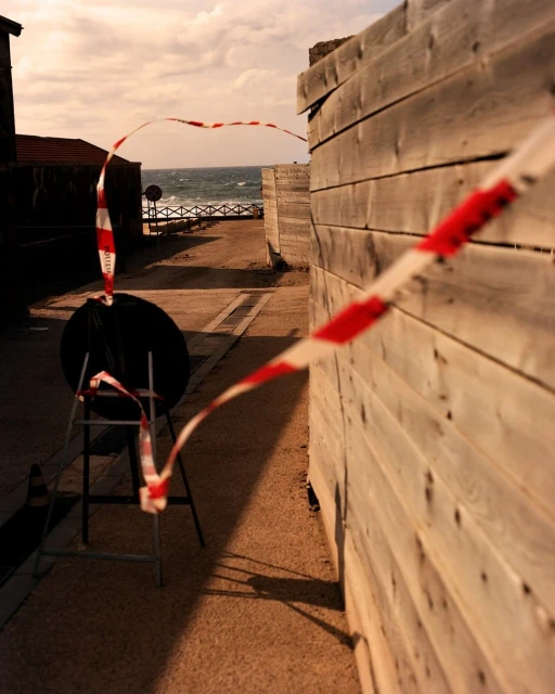 Coastal scene with a wooden wall and safety tape, leading to the ocean with a distant building, under a bright sky.