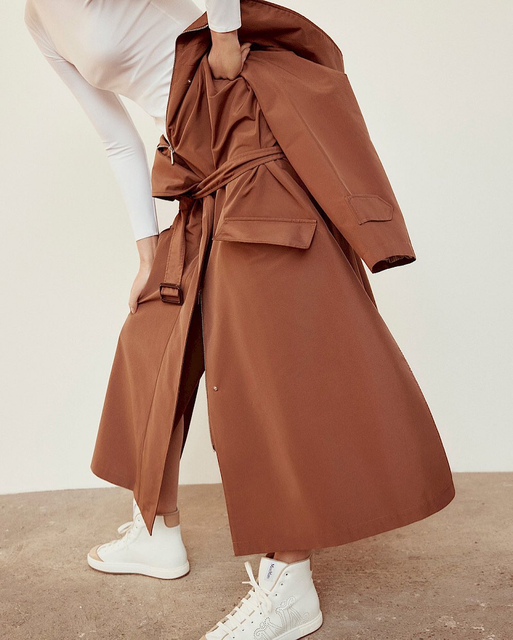 A brown trench coat is held by a person wearing white top, tights, and sneakers against a neutral backdrop in an indoor setting.
