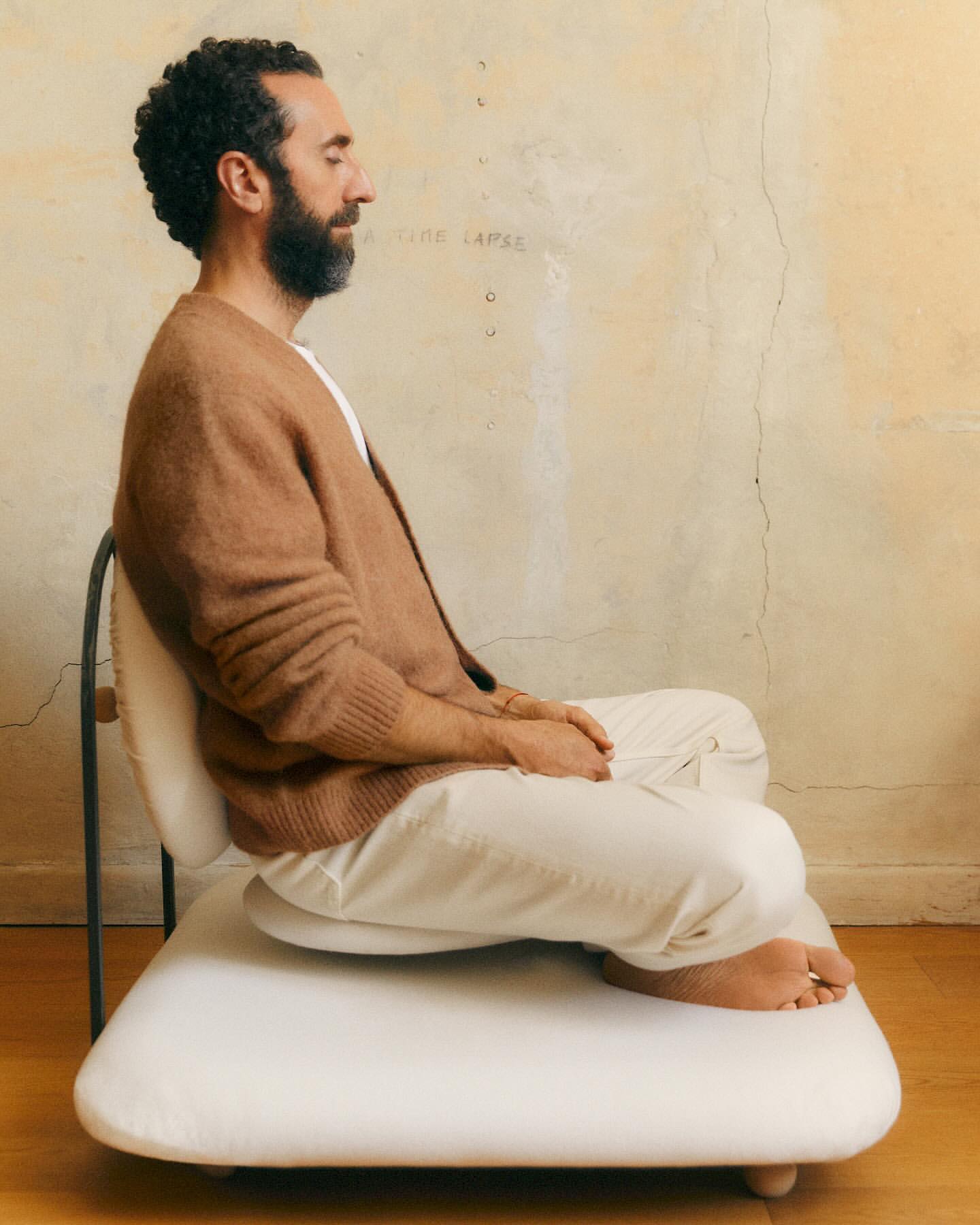 Adult male with a beard sits cross-legged on a padded chair in a room with a neutral-colored wall, looking relaxed.