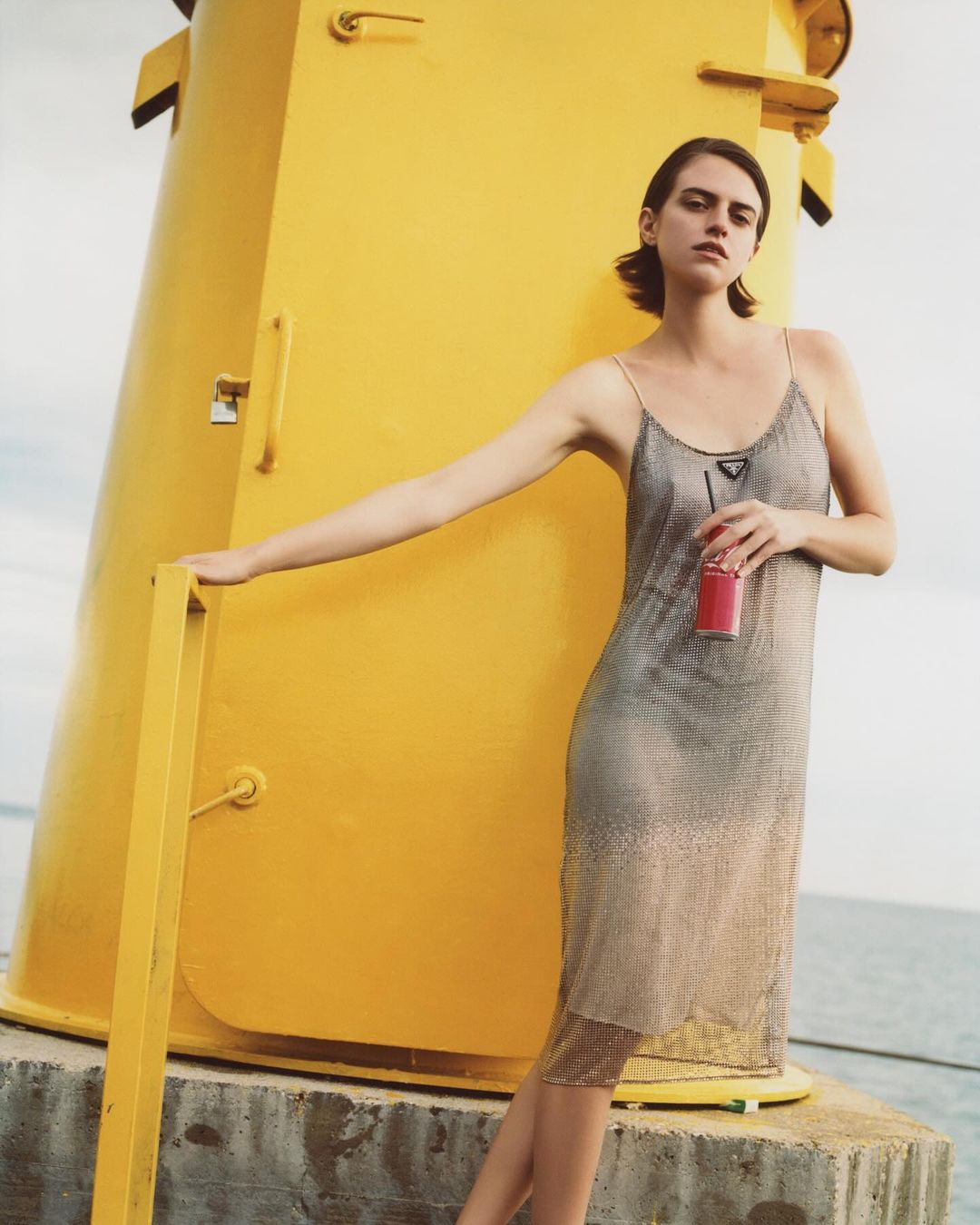 A young adult female stands holding a red soda can in a silver dress in front of a yellow lighthouse on a concrete base.