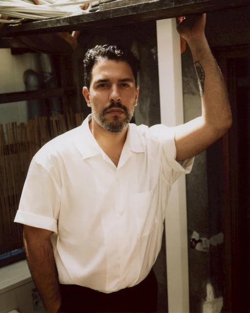 Portrait of a man with a beard, wearing a white button-down shirt, and posing outdoors with a neutral expression in natural light.
