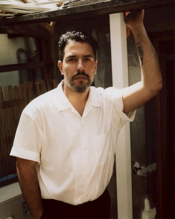 Portrait of a man with a beard, wearing a white button-down shirt, and posing outdoors with a neutral expression in natural light.