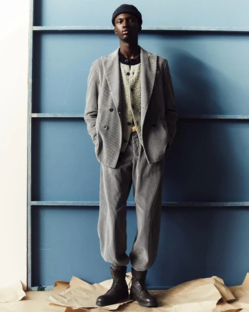 Full-length portrait of a Black man wearing a grey corduroy suit and dark beanie against a blue wall background.