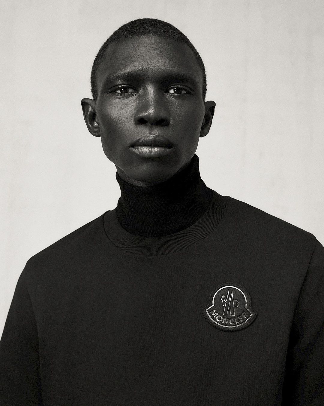 Portrait of a dark-skinned male fashion model in a black turtleneck and sweatshirt with Moncler logo against a plain gray background.