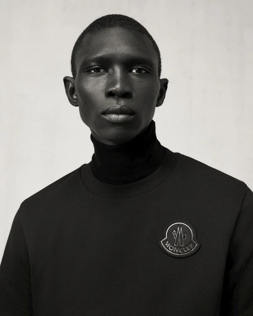 Portrait of a dark-skinned male fashion model in a black turtleneck and sweatshirt with Moncler logo against a plain gray background.
