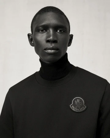 Portrait of a dark-skinned male fashion model in a black turtleneck and sweatshirt with Moncler logo against a plain gray background.