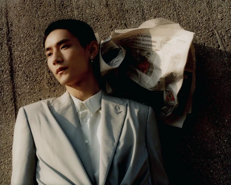 Portrait of a stylish man in a suit leaning against a textured wall with newspaper. Soft, natural lighting highlights the details.