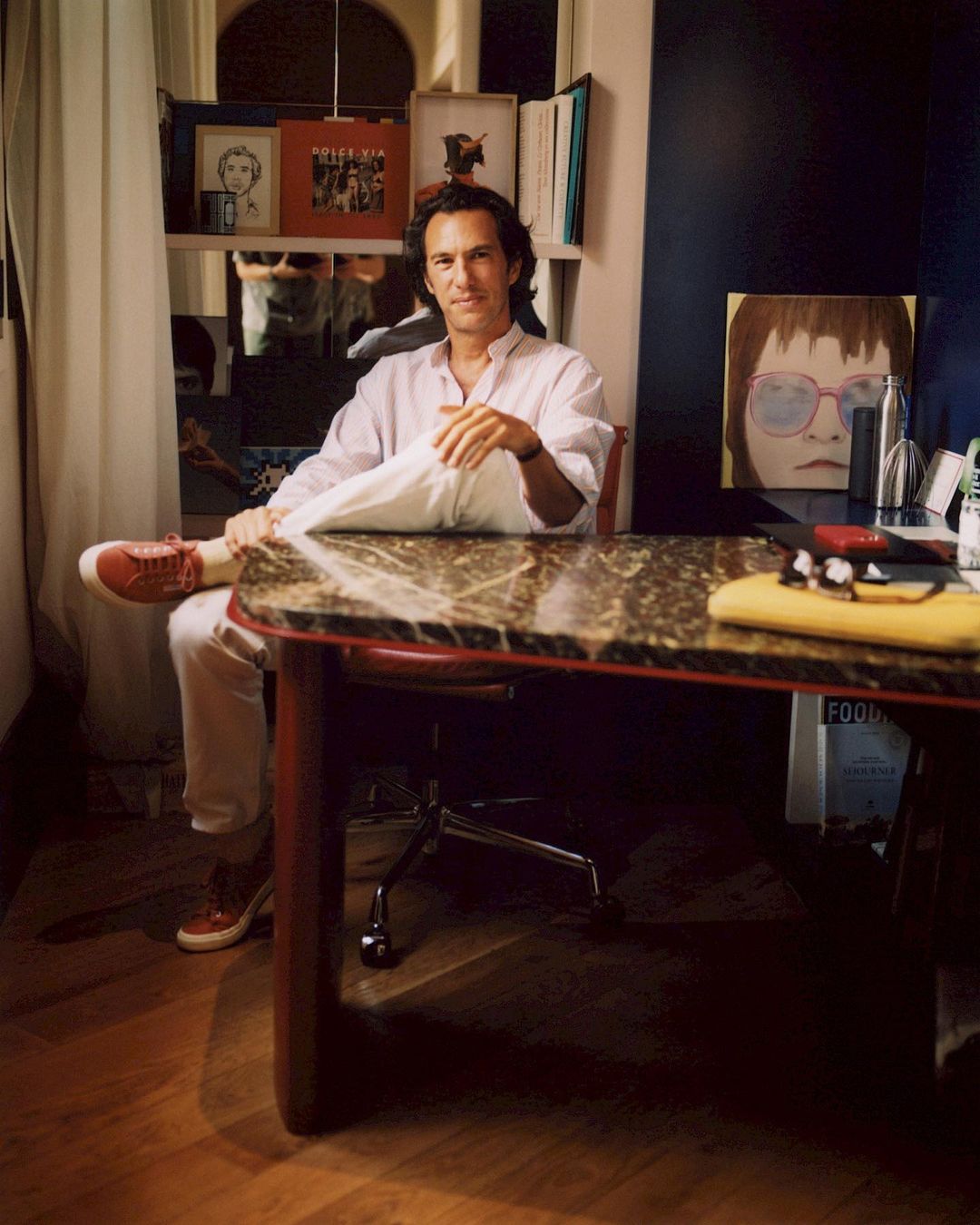 Adult male sitting at a marble table in a room with books and artwork, wearing a white shirt and red sneakers, creating an indoor scene.