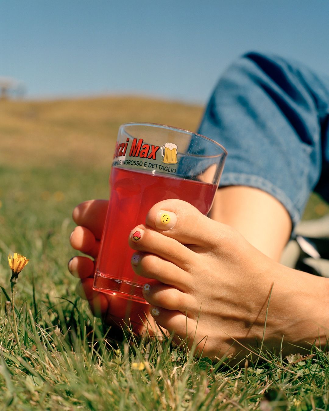 Close-up of adult bare feet with painted toenails holding a drink glass filled with red liquid, outdoors on a sunny day.