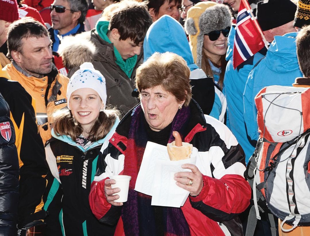 A crowd of people standing outdoors on a winter day; one woman in foreground eating a hot dog, other people in hats and jackets.