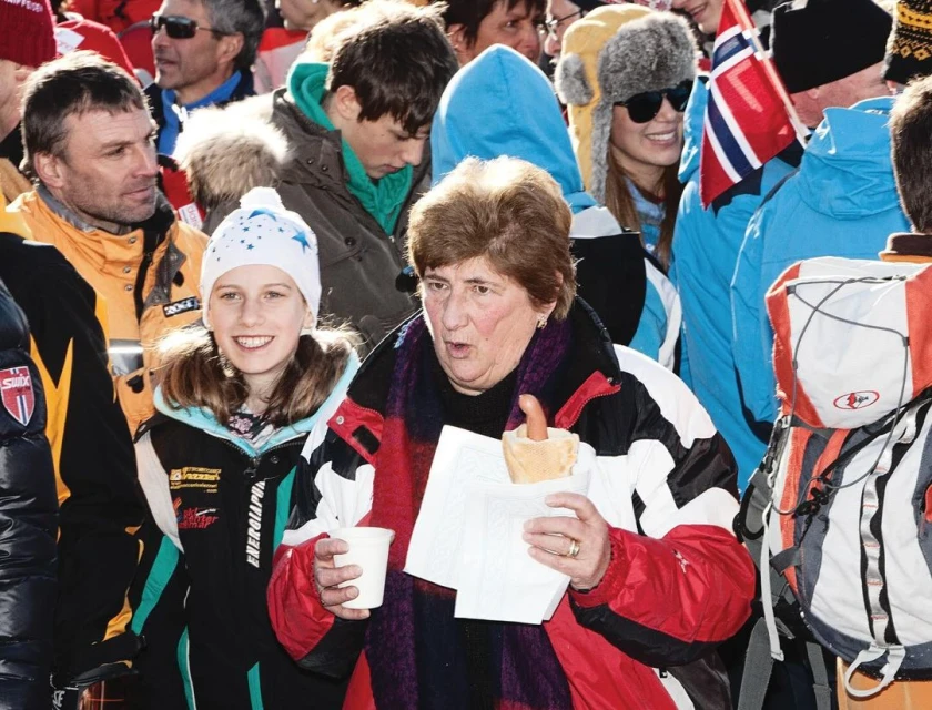 A crowd of people standing outdoors on a winter day; one woman in foreground eating a hot dog, other people in hats and jackets.