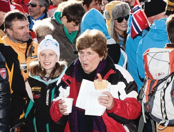 A crowd of people standing outdoors on a winter day; one woman in foreground eating a hot dog, other people in hats and jackets.