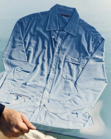 Blue long-sleeve button-down shirt laid flat on a reflective glass surface outdoors.