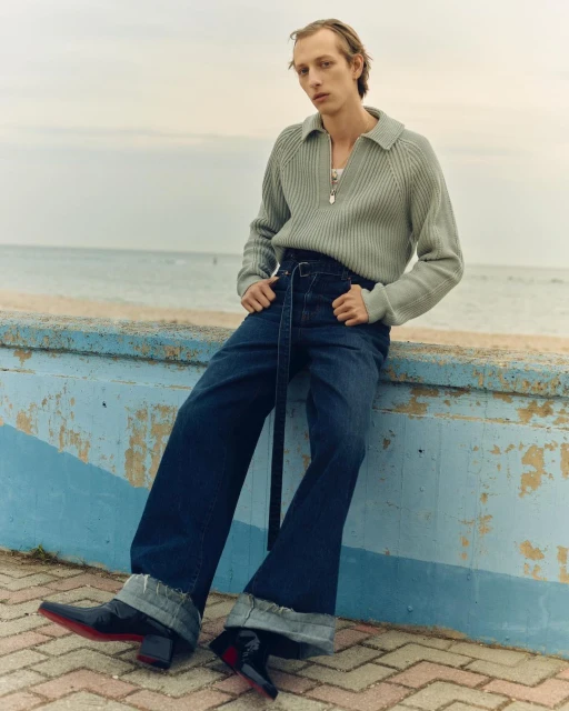 A slim, fair-skinned man in a grey sweater and wide-leg jeans poses outdoors on a blue wall in front of the beach on an overcast day.