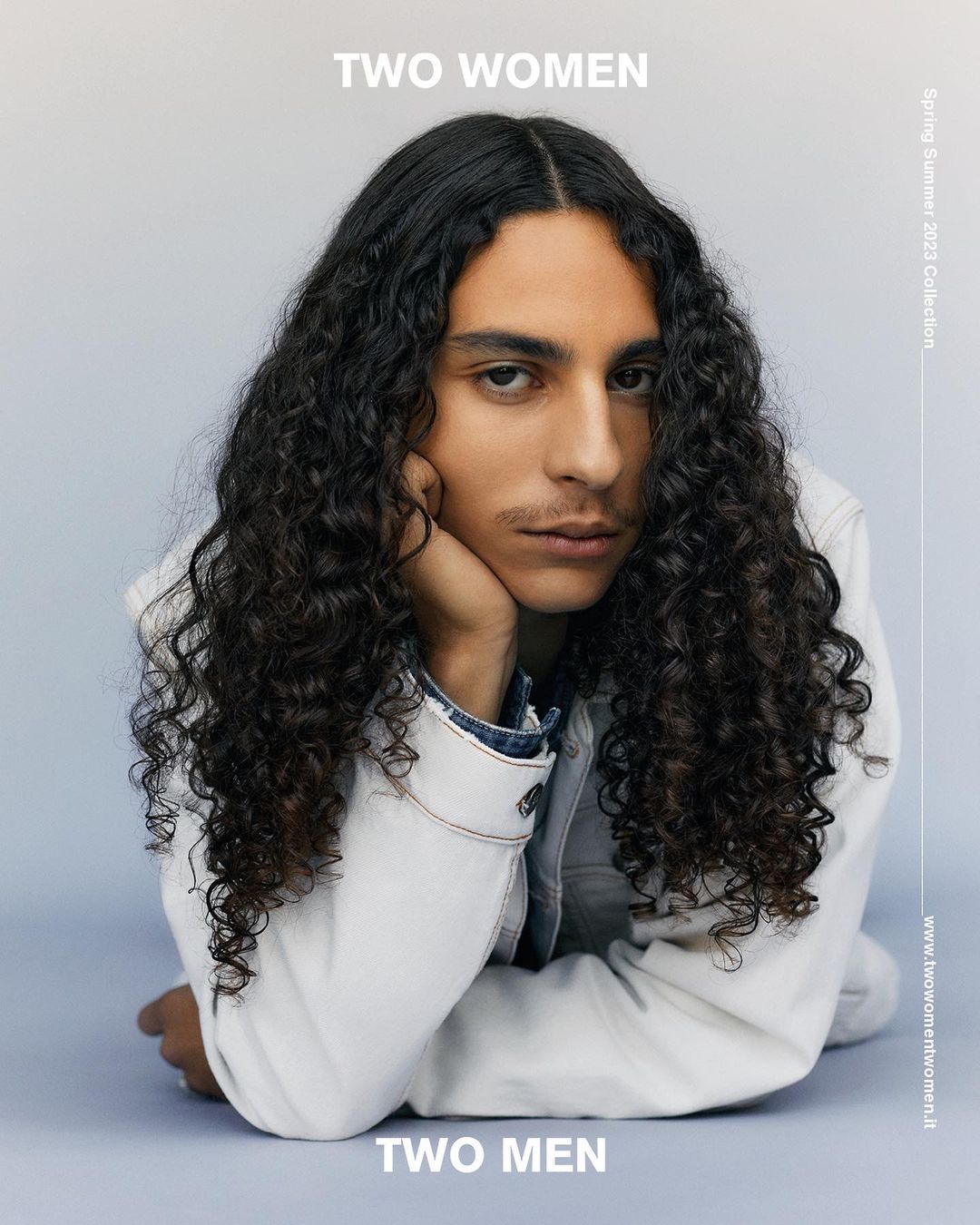 Portrait of an adult male with long curly hair, wearing a denim jacket, posing against a soft blue gradient background in a studio setting.