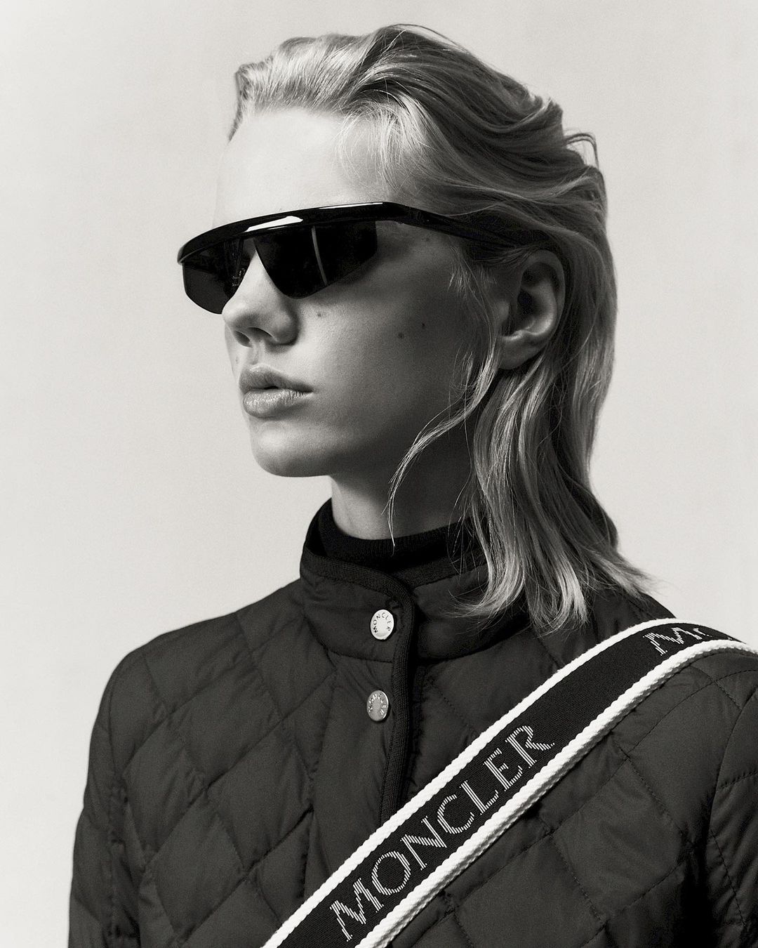 Monochrome portrait of a young woman with light blonde hair wearing sunglasses and a quilted jacket with a branded strap.
