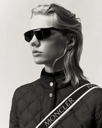 Monochrome portrait of a young woman with light blonde hair wearing sunglasses and a quilted jacket with a branded strap.