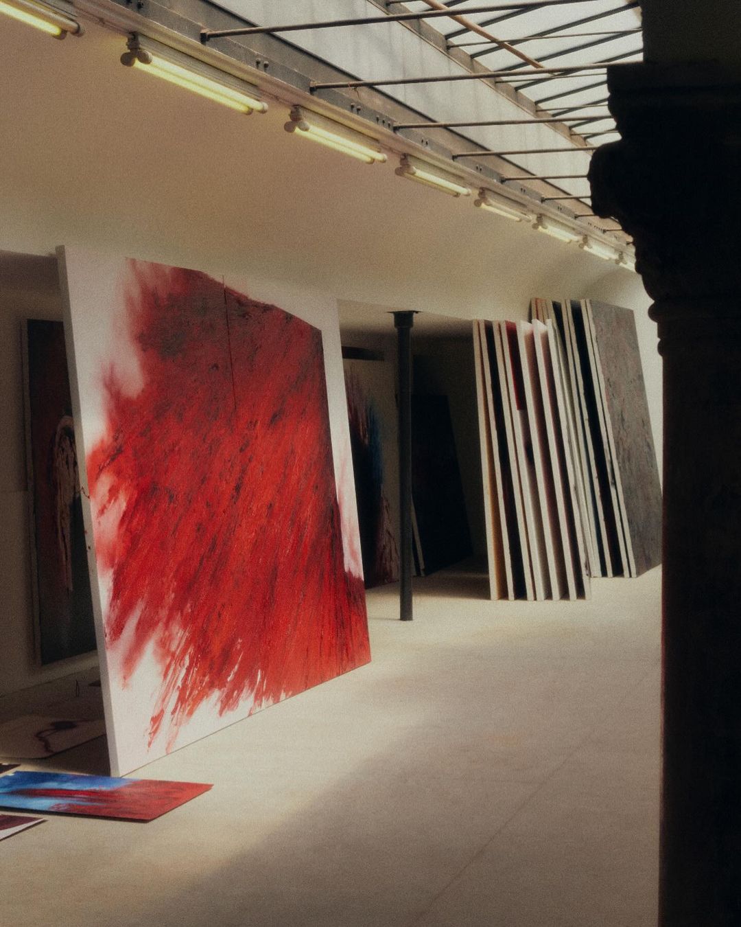 Large abstract paintings stand in a light-filled art studio with overhead fluorescent lighting and architectural column in the foreground.