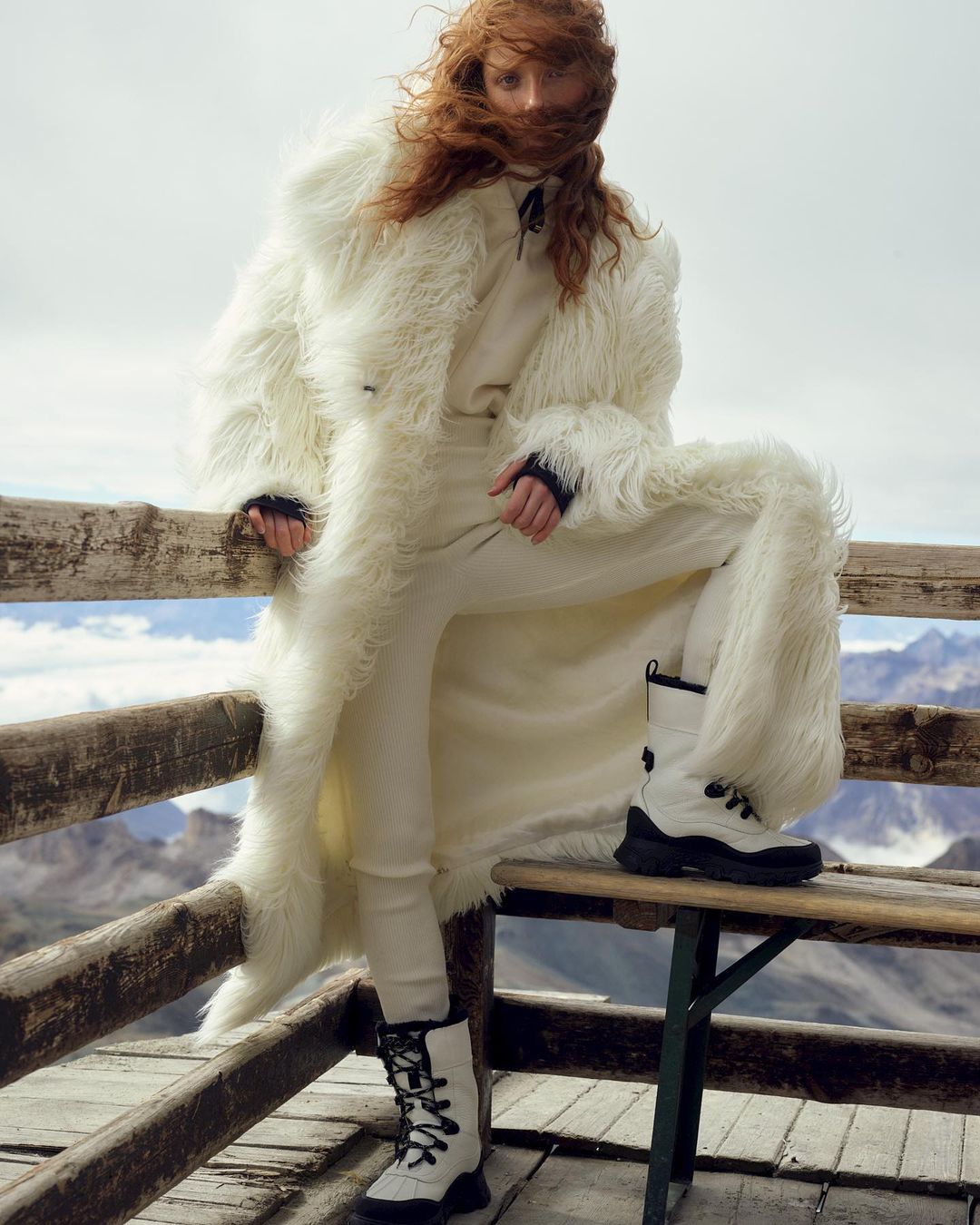 Woman in white faux-fur coat and boots poses on wooden structure in mountains. Red hair is windswept, off-white color palette.