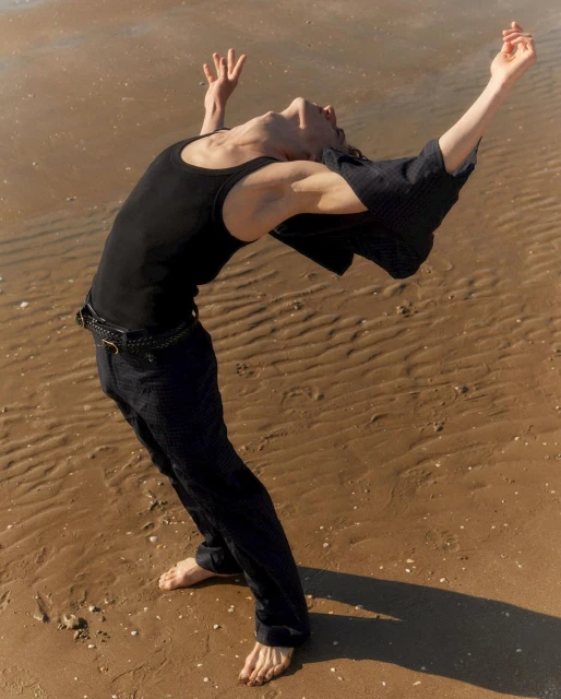 A person with dark hair and clothing bends backward on a sandy beach with arms raised. The individual is barefoot and facing upward.