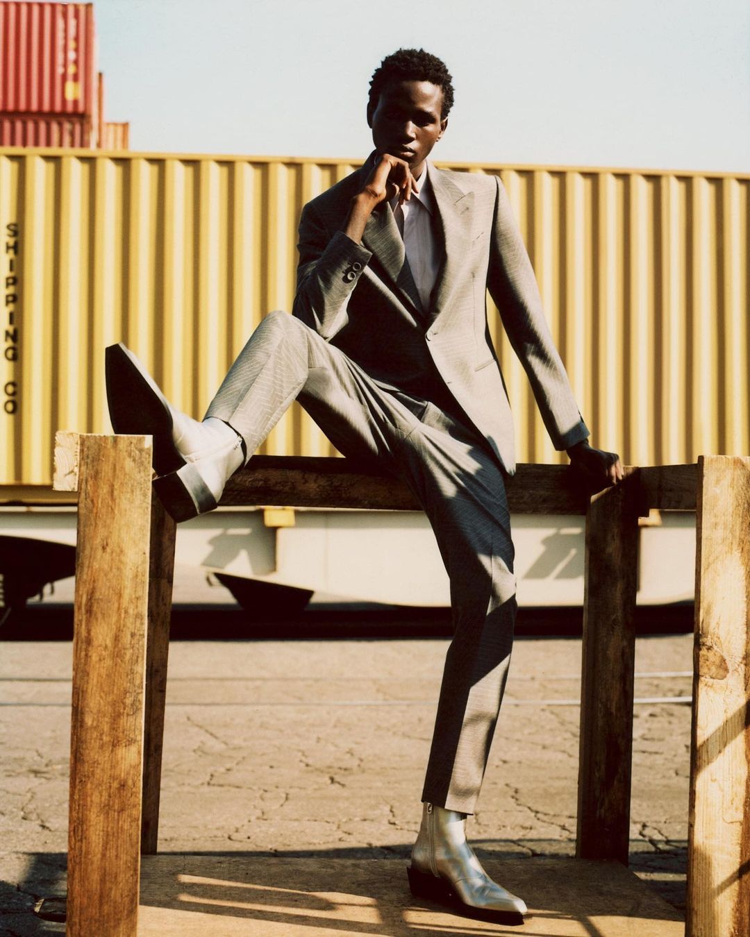 An adult Black male model in a gray suit poses on a wooden structure with shipping containers in the background for a fashion shoot.