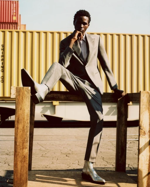 An adult Black male model in a gray suit poses on a wooden structure with shipping containers in the background for a fashion shoot.