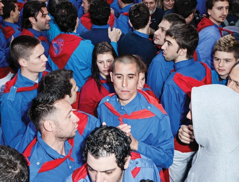Large group of adult males, most wearing matching blue collared shirts with red scarves, gathered indoors. Crowd of people. 