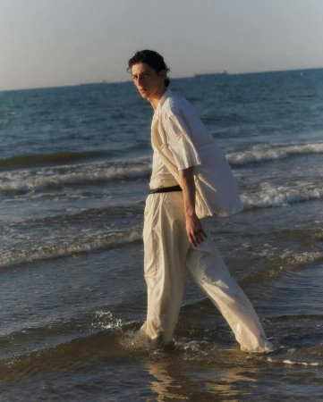 A young man in a white shirt and trousers walks along the beach in the water at daytime, with the blue sea and horizon behind him.