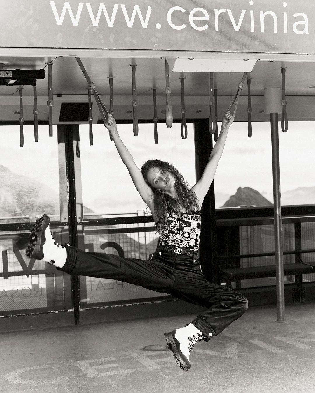 Black and white shot of a woman hanging from metal bars, wearing a Chanel shirt, pants, and boots with mountains in the background.