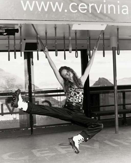 Black and white shot of a woman hanging from metal bars, wearing a Chanel shirt, pants, and boots with mountains in the background.
