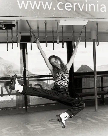 Black and white shot of a woman hanging from metal bars, wearing a Chanel shirt, pants, and boots with mountains in the background.