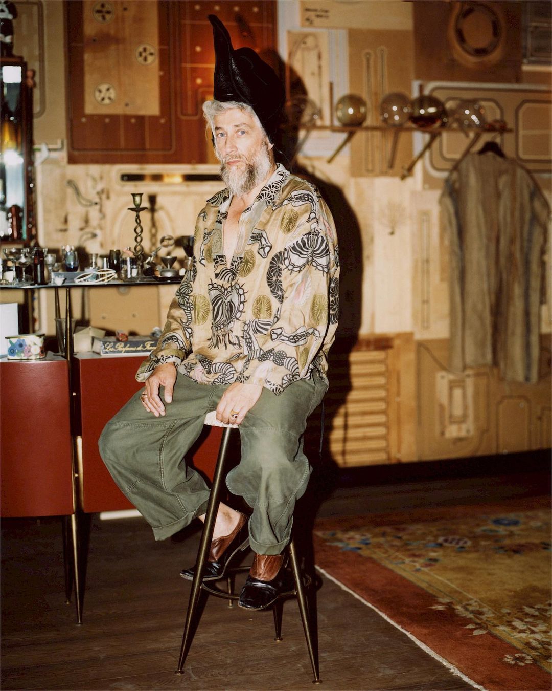 Older adult male with beard sits indoors wearing tunic-style shirt and pointy leather hat. Stool, table, vintage objects are visible.