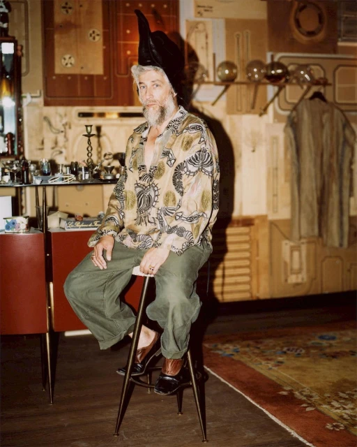 Older adult male with beard sits indoors wearing tunic-style shirt and pointy leather hat. Stool, table, vintage objects are visible.