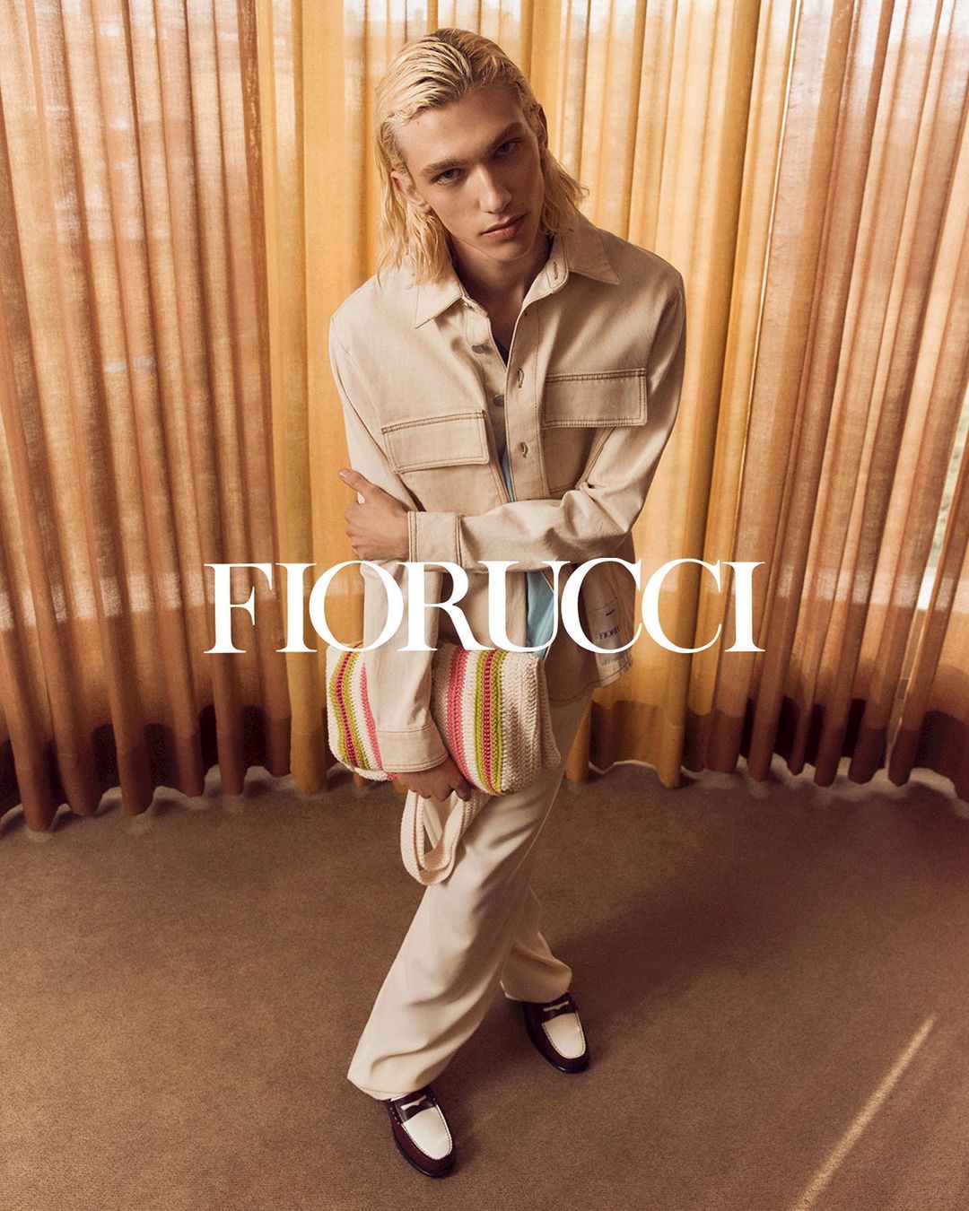 Indoor studio shot of a blond male model posing in a neutral outfit with a multicolored woven handbag from fashion brand Fiorucci.