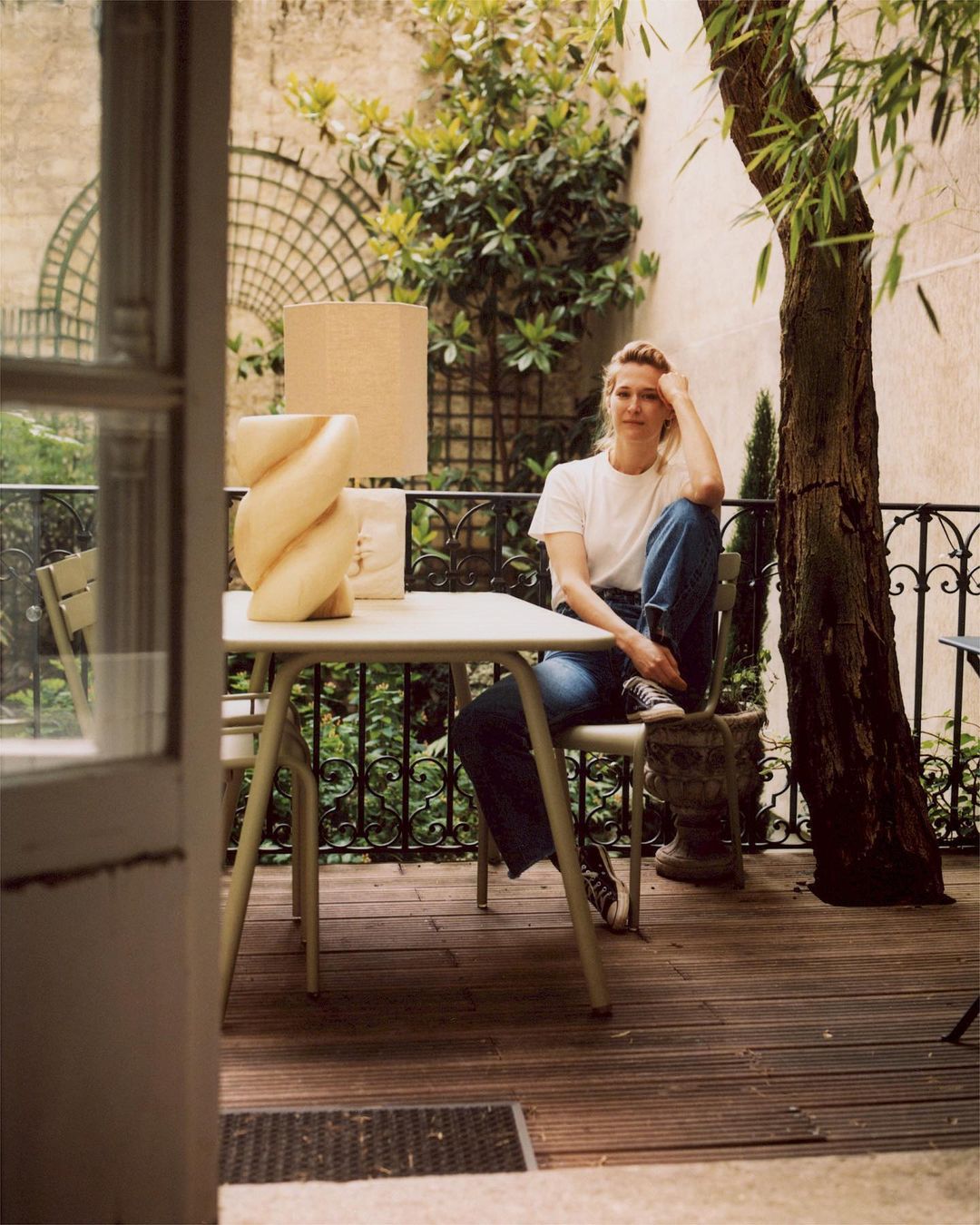 Adult woman with blond hair is sitting at a table with a lamp in an outdoor patio setting, surrounded by greenery and trees.