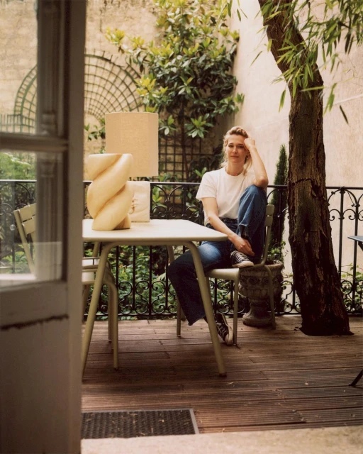 Adult woman with blond hair is sitting at a table with a lamp in an outdoor patio setting, surrounded by greenery and trees.