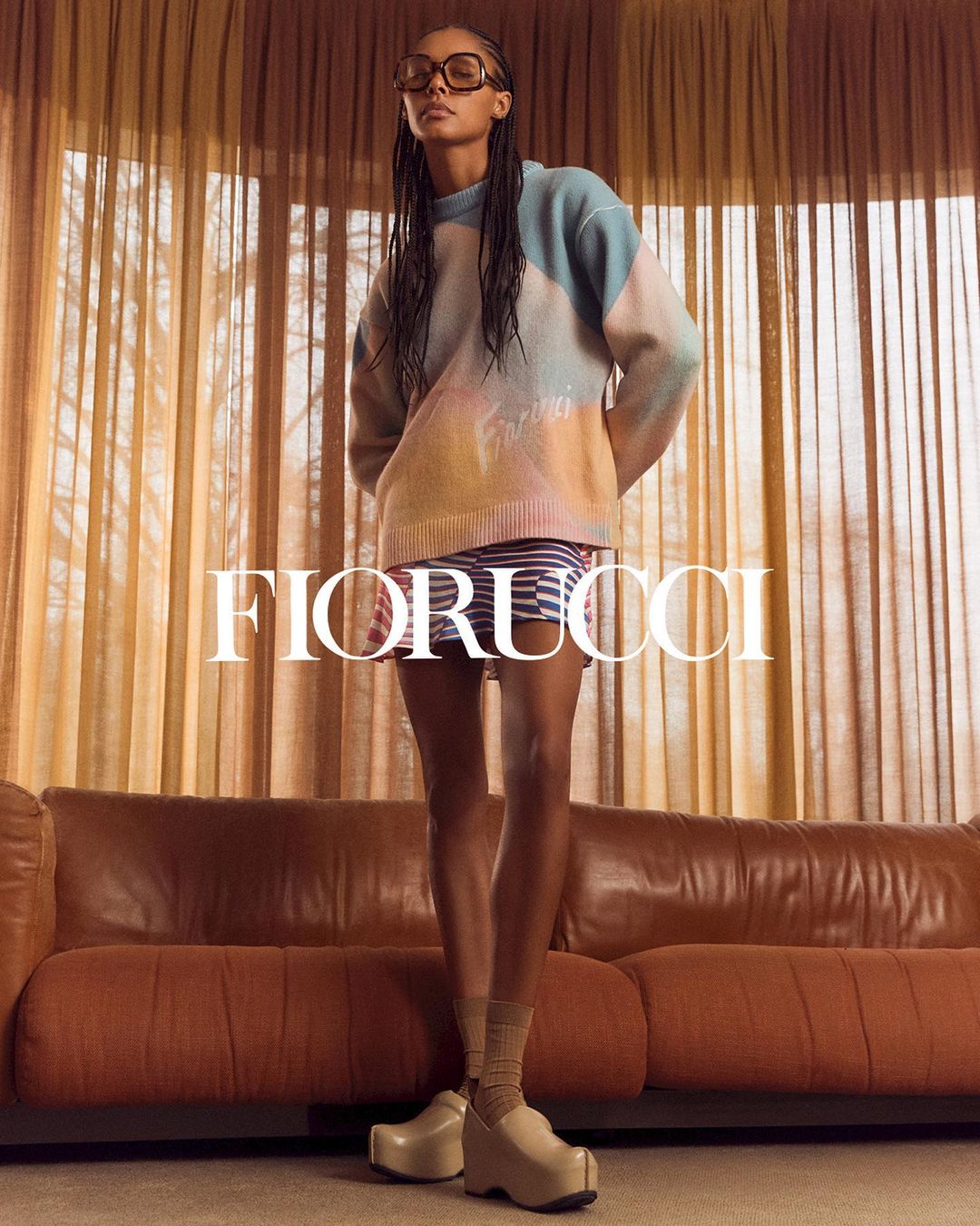 A young adult female stands in front of curtains wearing a colorful sweater, striped skirt, platform shoes, and glasses in a studio setting.