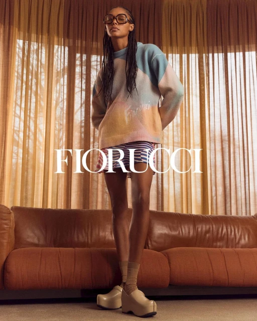 A young adult female stands in front of curtains wearing a colorful sweater, striped skirt, platform shoes, and glasses in a studio setting.