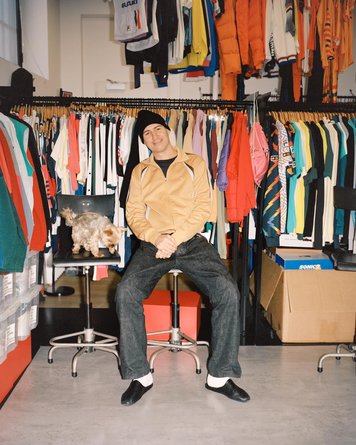 A man sits on a red stool in a clothing store, a small dog sits on a chair behind him. Racks of clothing line the walls.