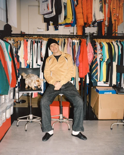 A man sits on a red stool in a clothing store, a small dog sits on a chair behind him. Racks of clothing line the walls.
