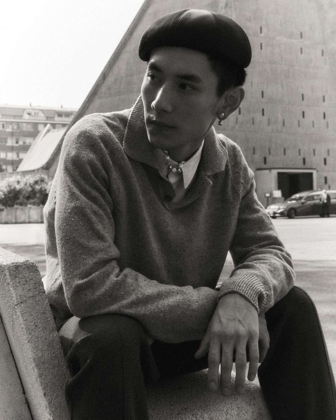 Portrait of a young Asian man wearing a black beret and sweater, sitting outside on a concrete structure near a modern building.