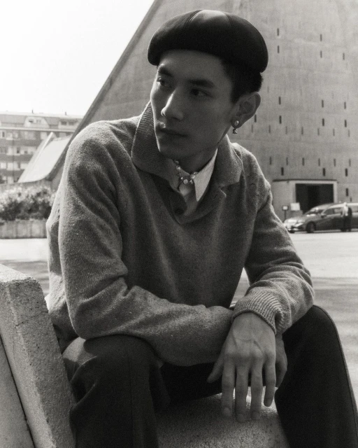 Portrait of a young Asian man wearing a black beret and sweater, sitting outside on a concrete structure near a modern building.
